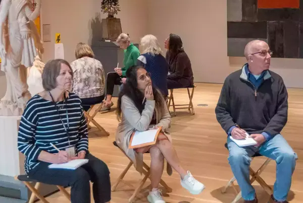 Museum visitors sit on folding stools, sketching and taking notes in the Snite Museum of Art.