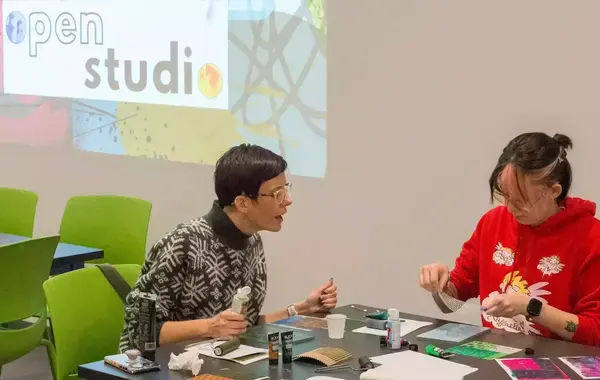 Two people work on art projects at a table.  An "Open Studio" logo is projected on the wall.  One person, wearing glasses and a patterned sweater, gestures while holding a tube of paint.  The other person, wearing a red hoodie, works with a piece of mesh material.  Art supplies are spread across the table.